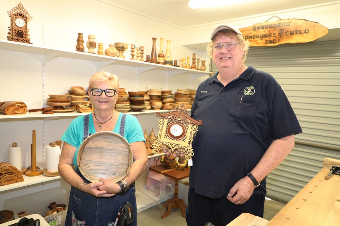 Twee personen in een moderne houtwerkplaats tonen handgemaakte houten voorwerpen, met een bord van de 'Bundaberg Woodworkers Guild Inc.' zichtbaar.